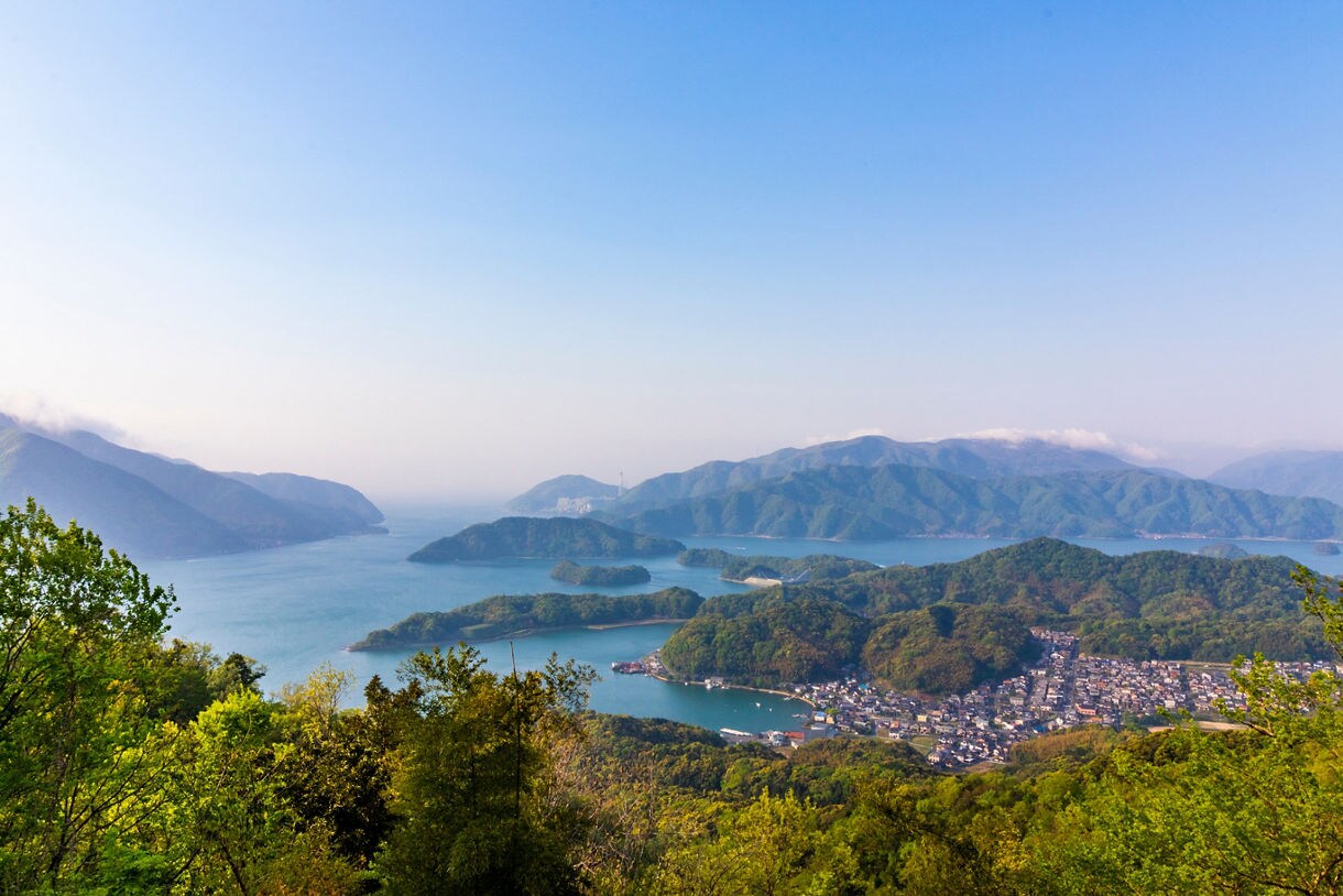 A wide scenic view of Maizuru Bay with clusters of green forested islands, blue water, and distant mountain ranges under a clear sky, with a small coastal town visible in the lower right.