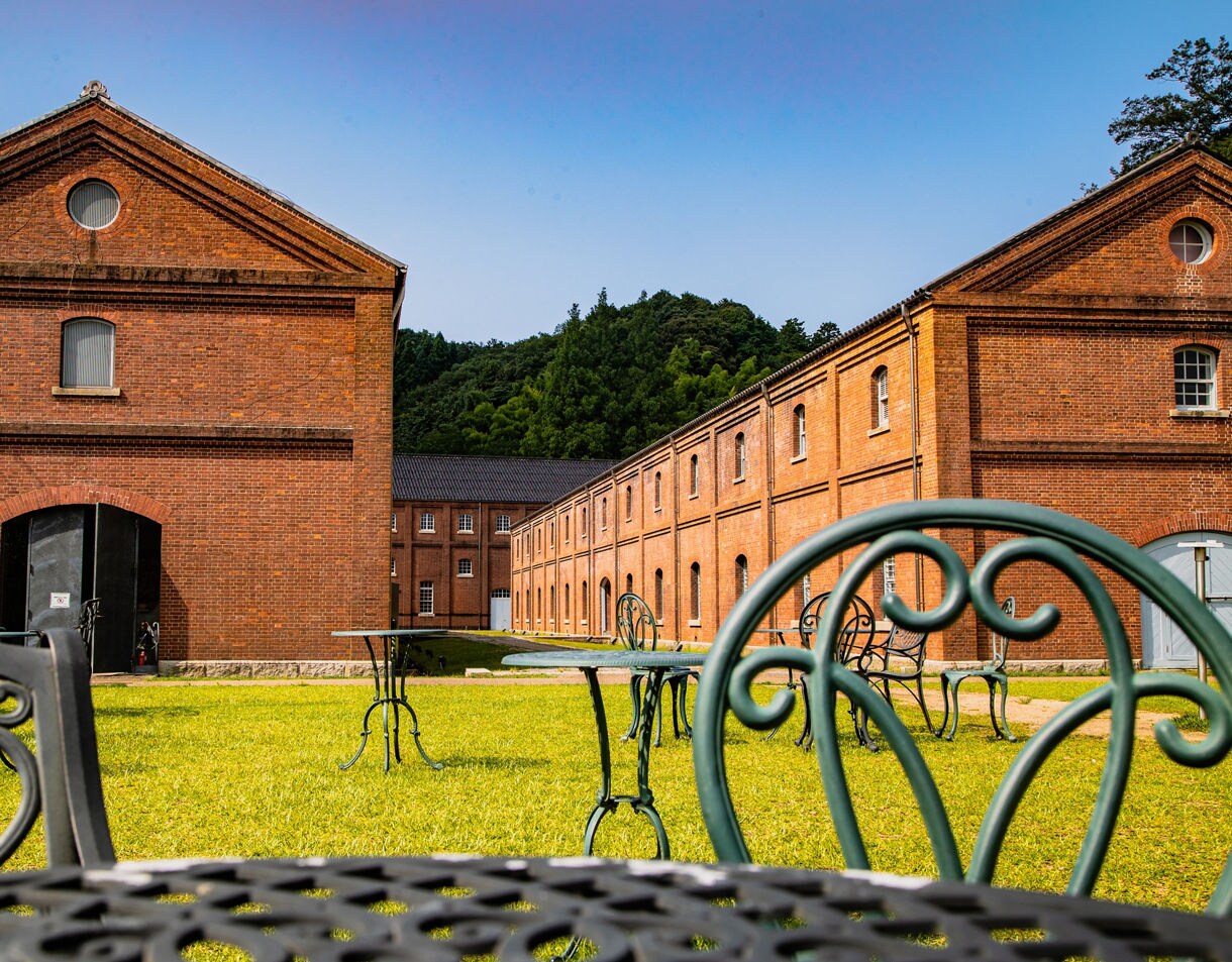 The photo shows Maizuru Red Brick Park with two long red brick warehouse buildings facing each other across a grassy courtyard. Metal café tables and chairs sit on the lawn, and forested hills rise behind the buildings under a clear blue sky.