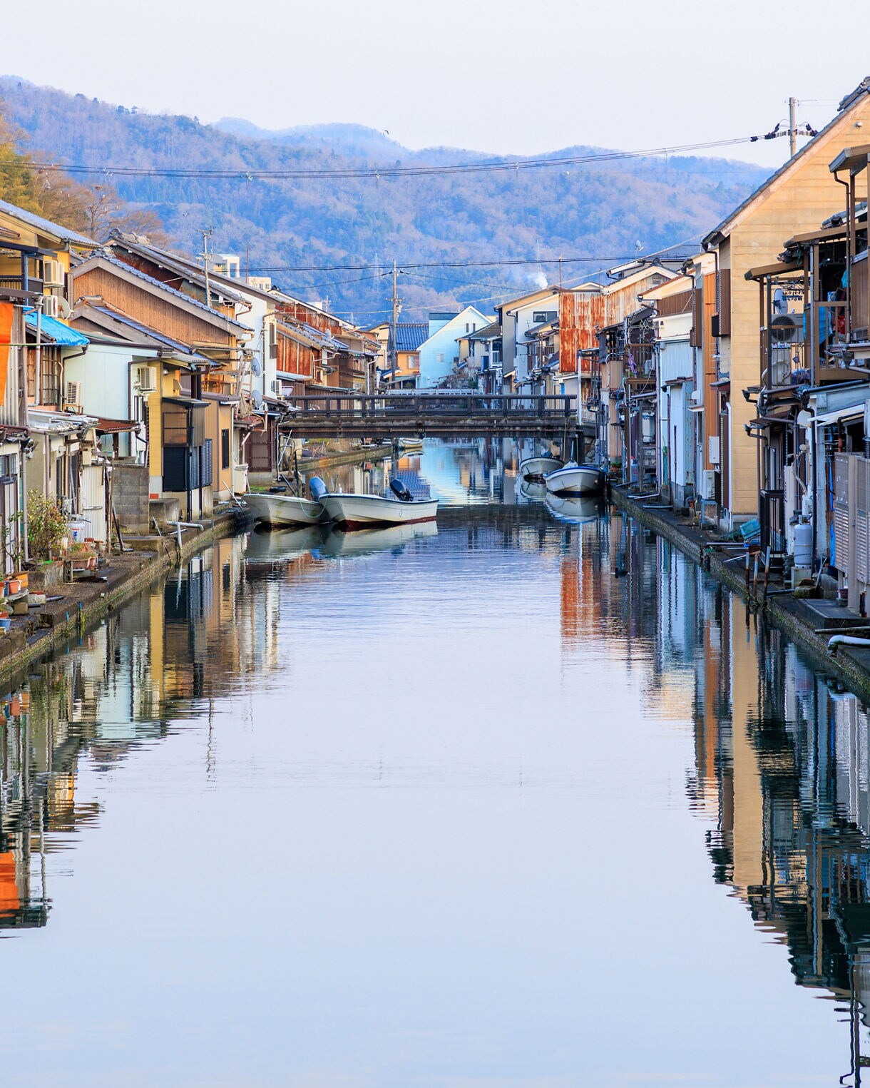 A quiet canal lined with traditional wooden houses in Maizuru, Japan, with small boats moored along the water and distant blue mountains rising in the background. The calm surface of the canal reflects the homes and sky.