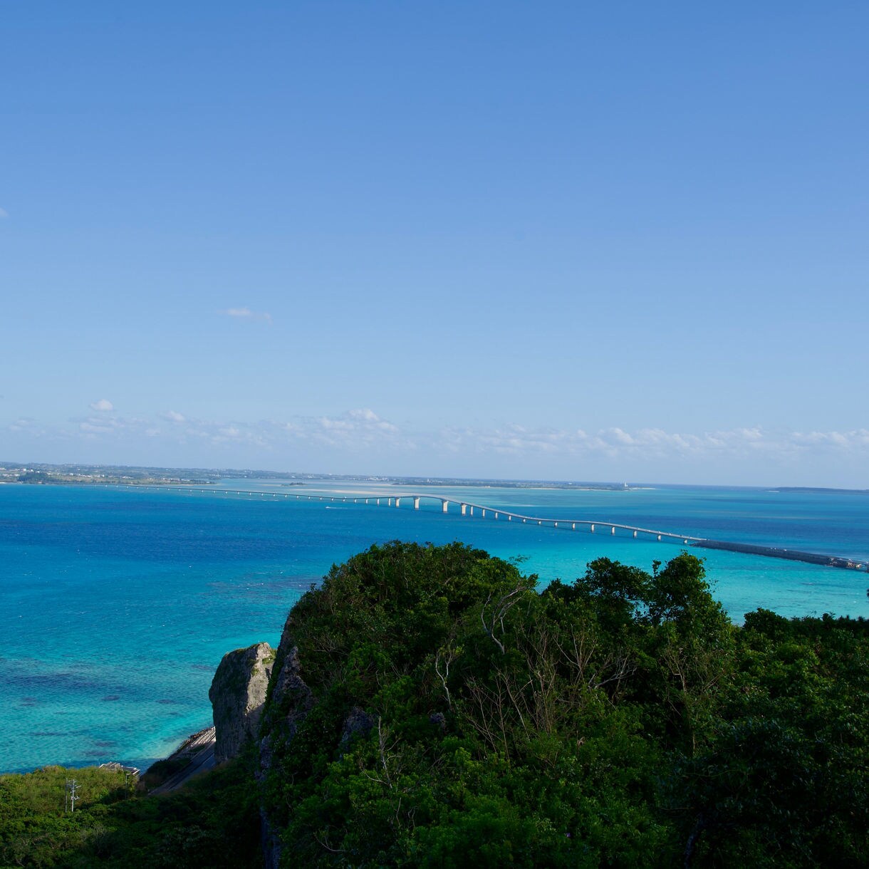 Elevated view from Makiyama Observatory overlooking vivid turquoise seas, lush green cliffs and a long bridge curving across the water toward distant islands under a clear blue sky.