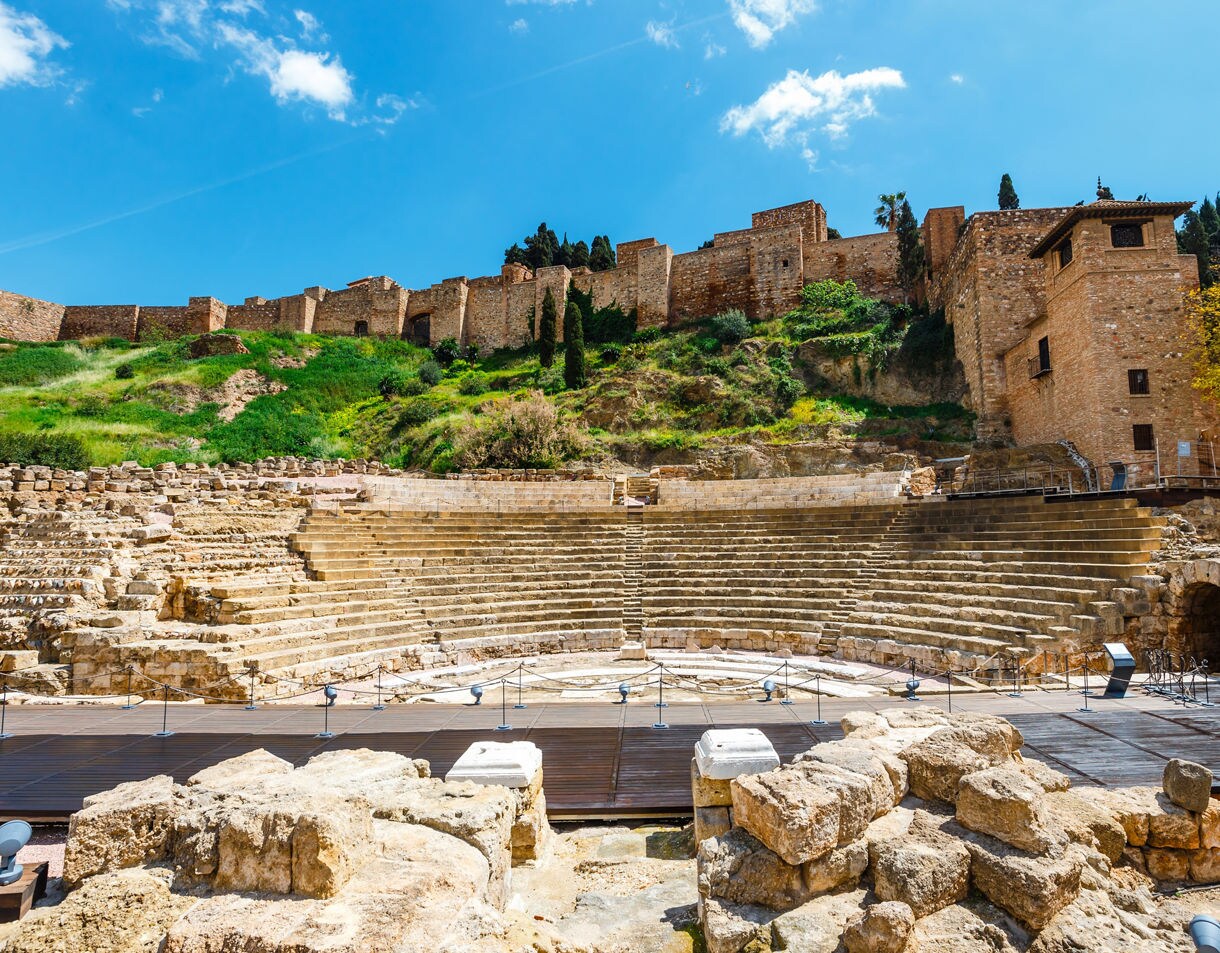 Ancient Roman Theatre in Málaga with semicircular stone seating, overlooked by Alcazaba fortress walls on the hillside.