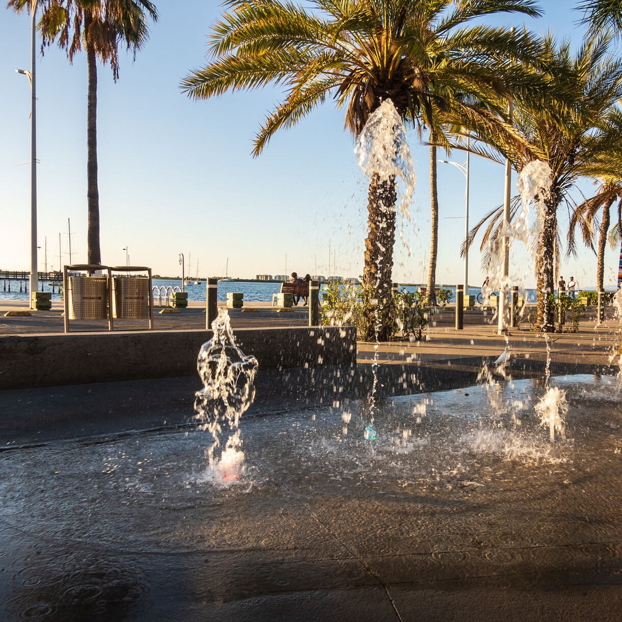 Water fountains spraying along the malecón in La Paz, framed by palm trees, ocean views and a mural with a Frida Kahlo design.