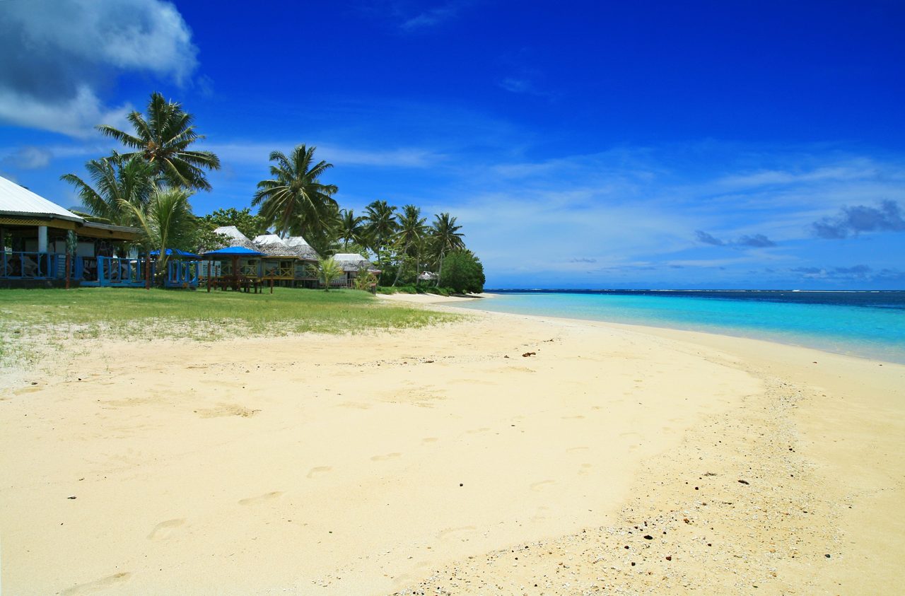 A tranquil stretch of Manase Beach with soft golden sand, turquoise ocean and palm trees lining beachfront bungalows.