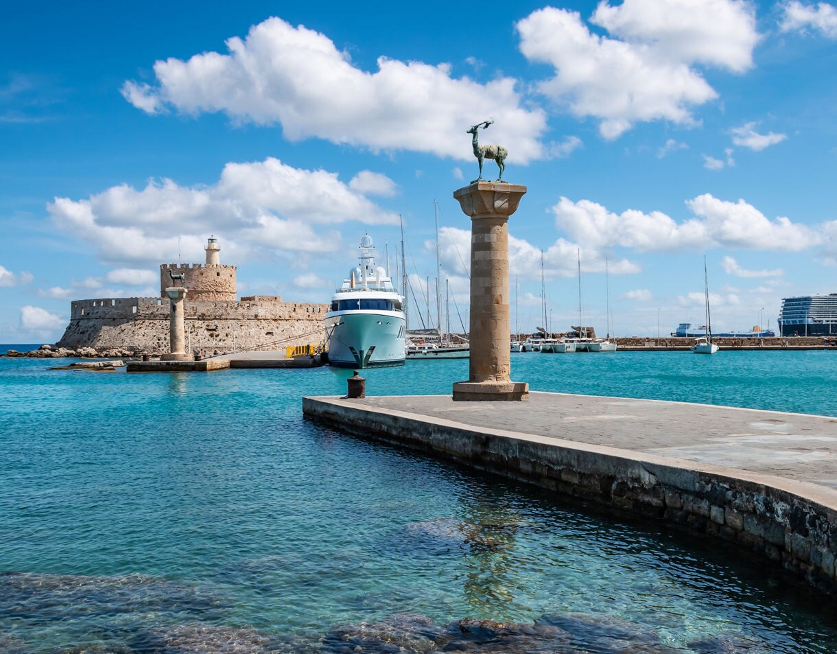 Mandraki Harbor in Rhodes, Greece, featuring stone deer statues on columns, a medieval fortress, yachts and turquoise waters under a sunny sky.