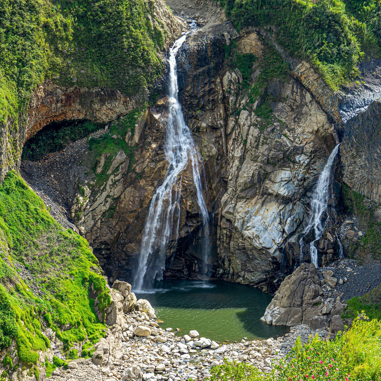 A tall double waterfall drops down rocky cliffs into a green pool surrounded by lush vegetation and scattered boulders.