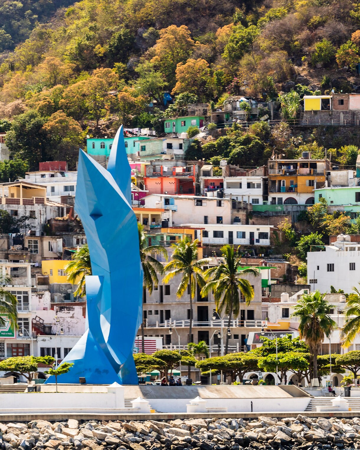 Vibrant hillside houses and the blue sailfish monument in downtown Manzanillo with palm trees and waterfront promenade.