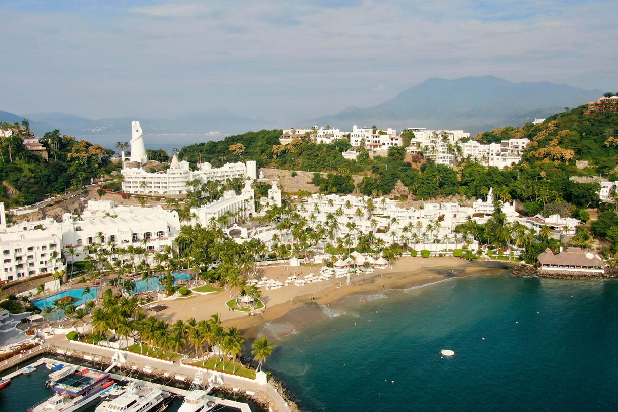 Aerial view of Manzanillo, Mexico with white beachfront resorts, palm-lined sands, a marina with yachts and green hills in the background.