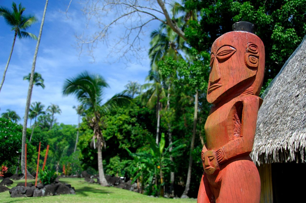 Red wooden Polynesian statue at Marae Arahurahu with tropical trees, palms and a thatched structure in the background.
