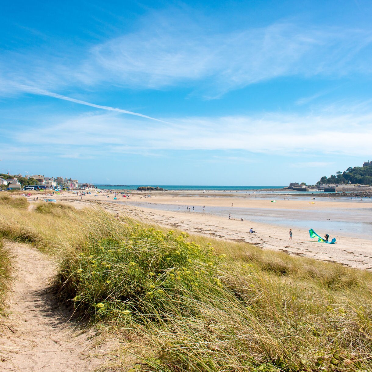 Sandy beach at Marazion with grassy dunes in the foreground, people walking along the shore and St Michael’s Mount rising on a small tidal island in the distance.