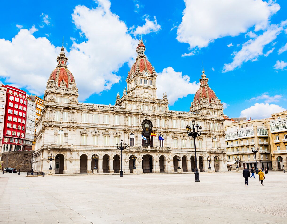 Wide plaza in A Coruña with the ornate City Hall building featuring three red-domed towers, arched walkways and surrounding colorful apartment facades under a bright blue sky.