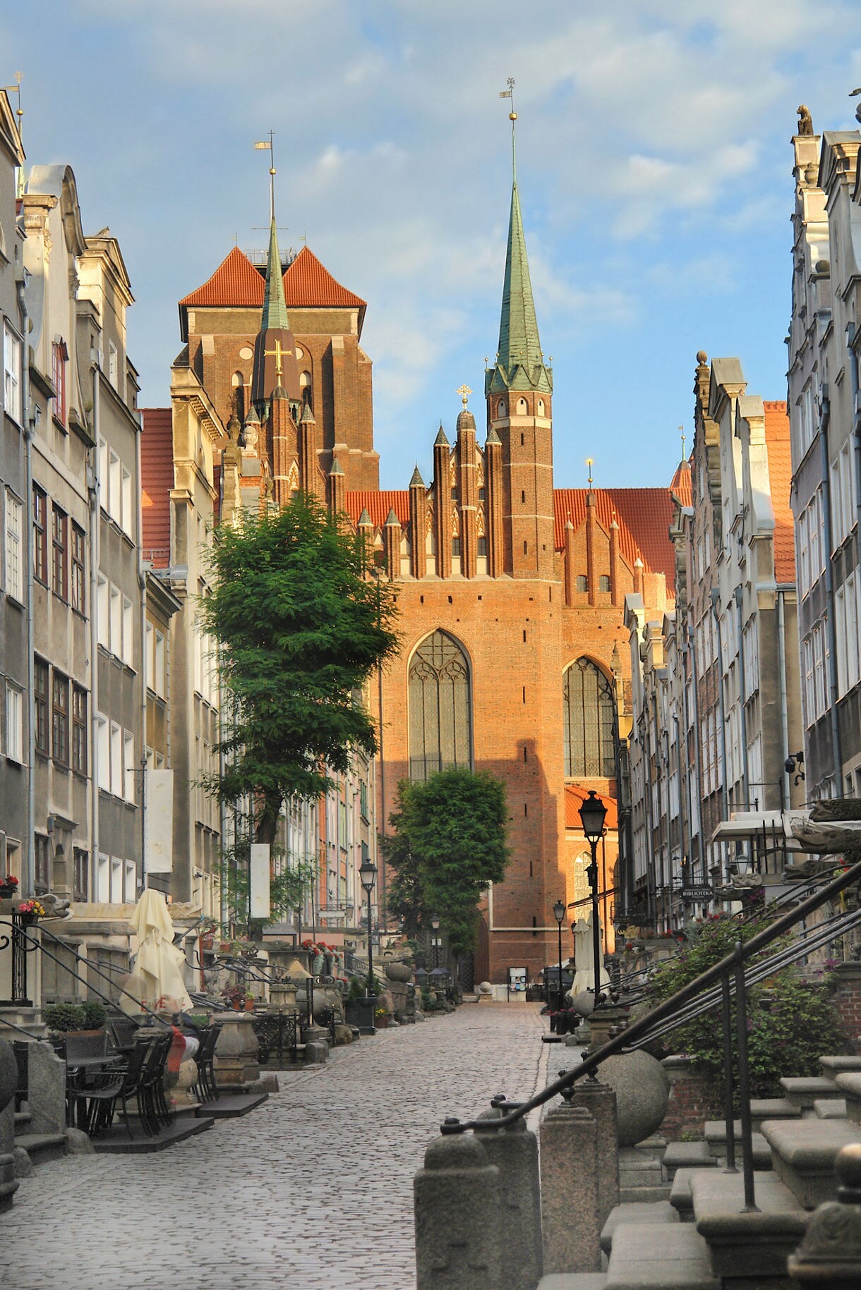Cobblestone Mariacka Street in Gdańsk with outdoor terraces, historic townhouses and St. Mary’s Basilica towering at the end of the street.
