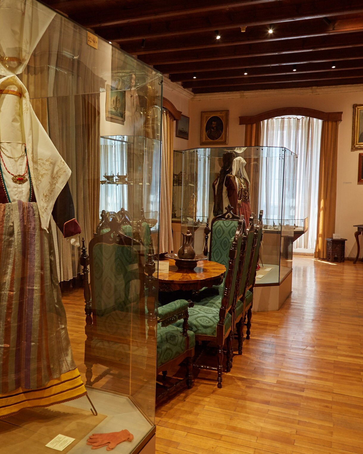 Display of Montenegrin traditional clothing in glass cases at the Maritime Museum of Kotor, with embroidered dresses, wooden furniture and framed portraits in a historic room.