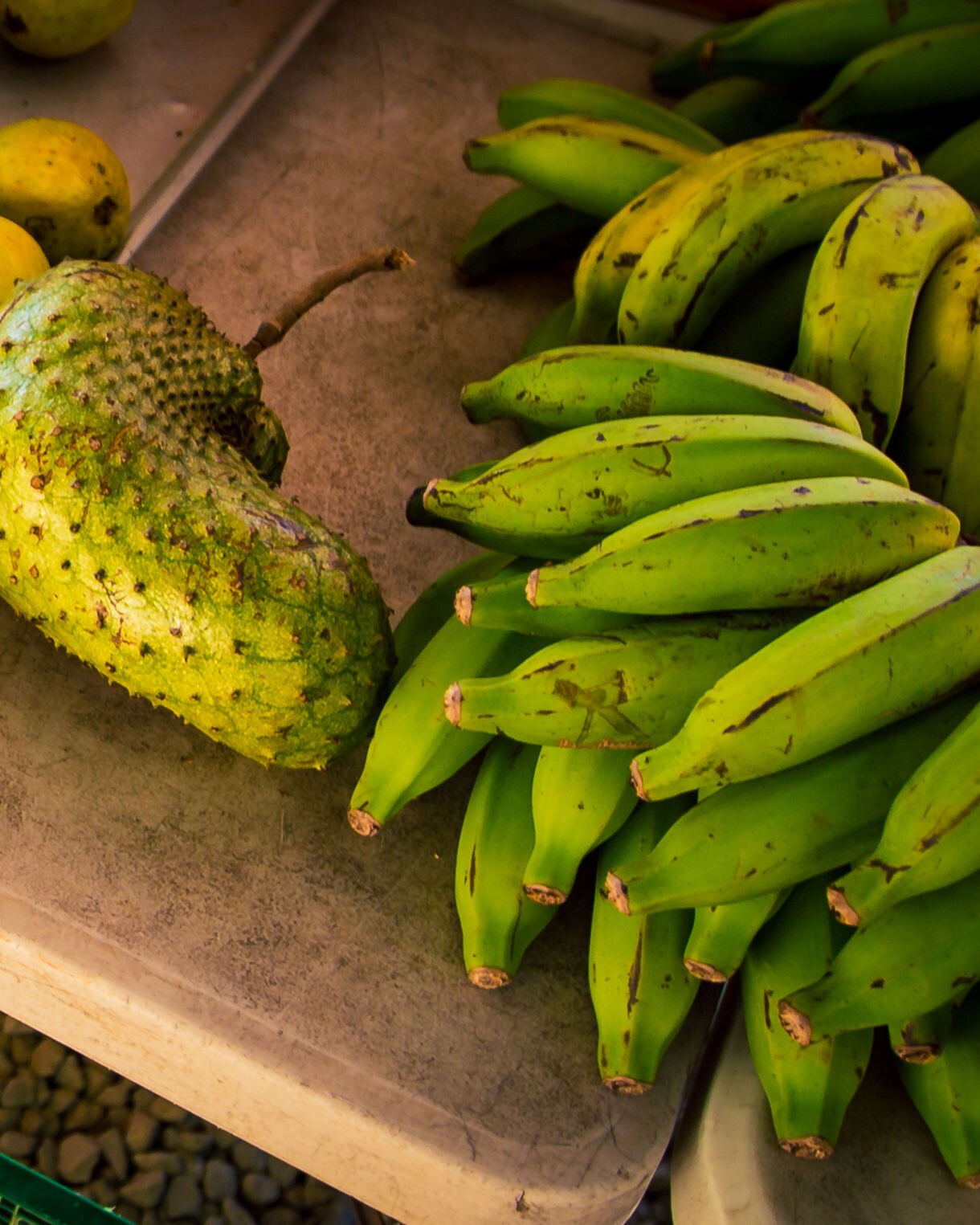Market stall display featuring piles of fresh tropical fruit including yellow guavas, green bananas, red rambutans and a large soursop.
