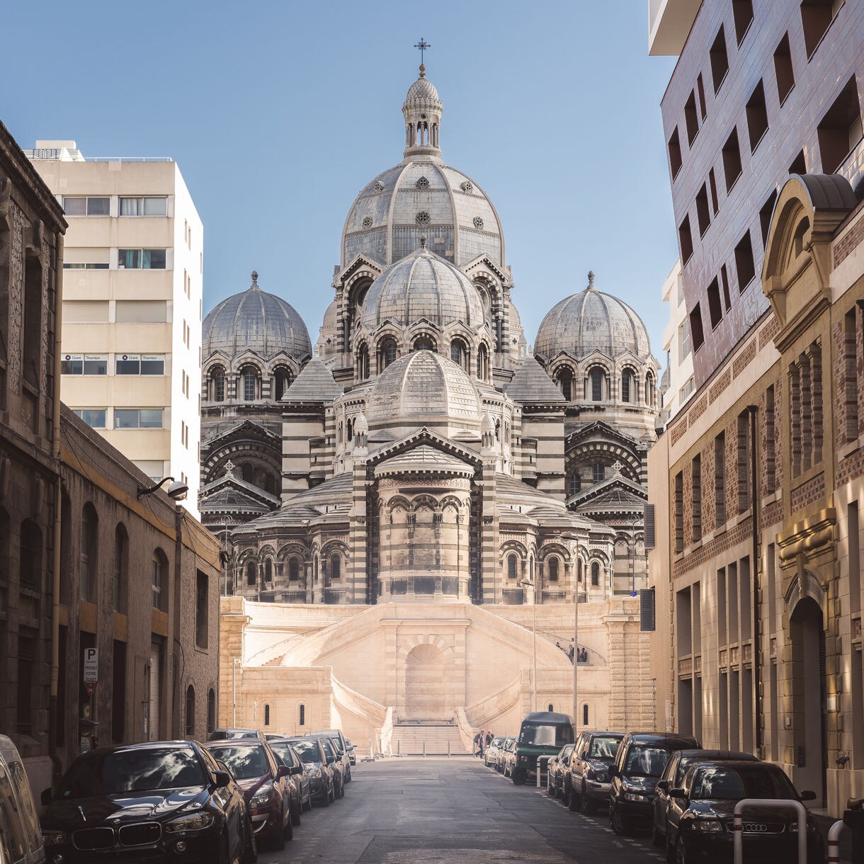 Street view in Marseille with parked cars leading toward the ornate domes and striped façade of Cathédrale de la Major.