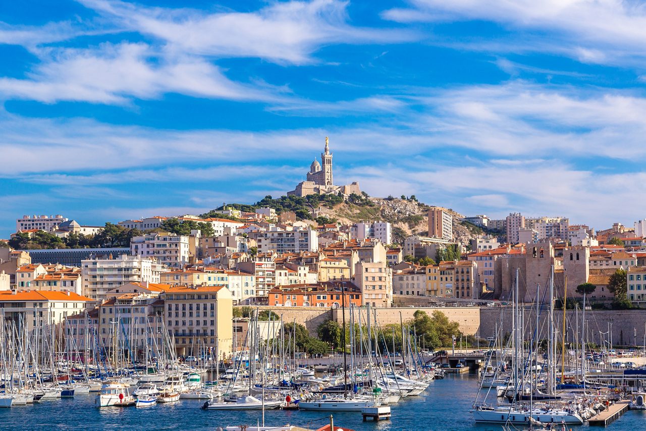 View of Marseille’s Old Port filled with sailboats, with the hilltop basilica of Notre-Dame de la Garde rising above the city under a vivid blue sky.