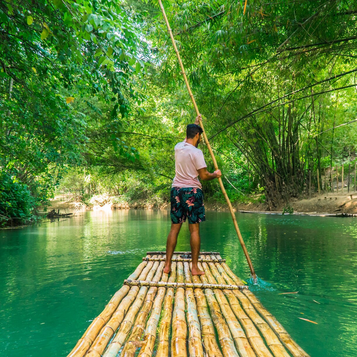 A man navigates a bamboo raft with a long pole through the calm, emerald waters of the Martha Brae River, framed by dense green foliage.