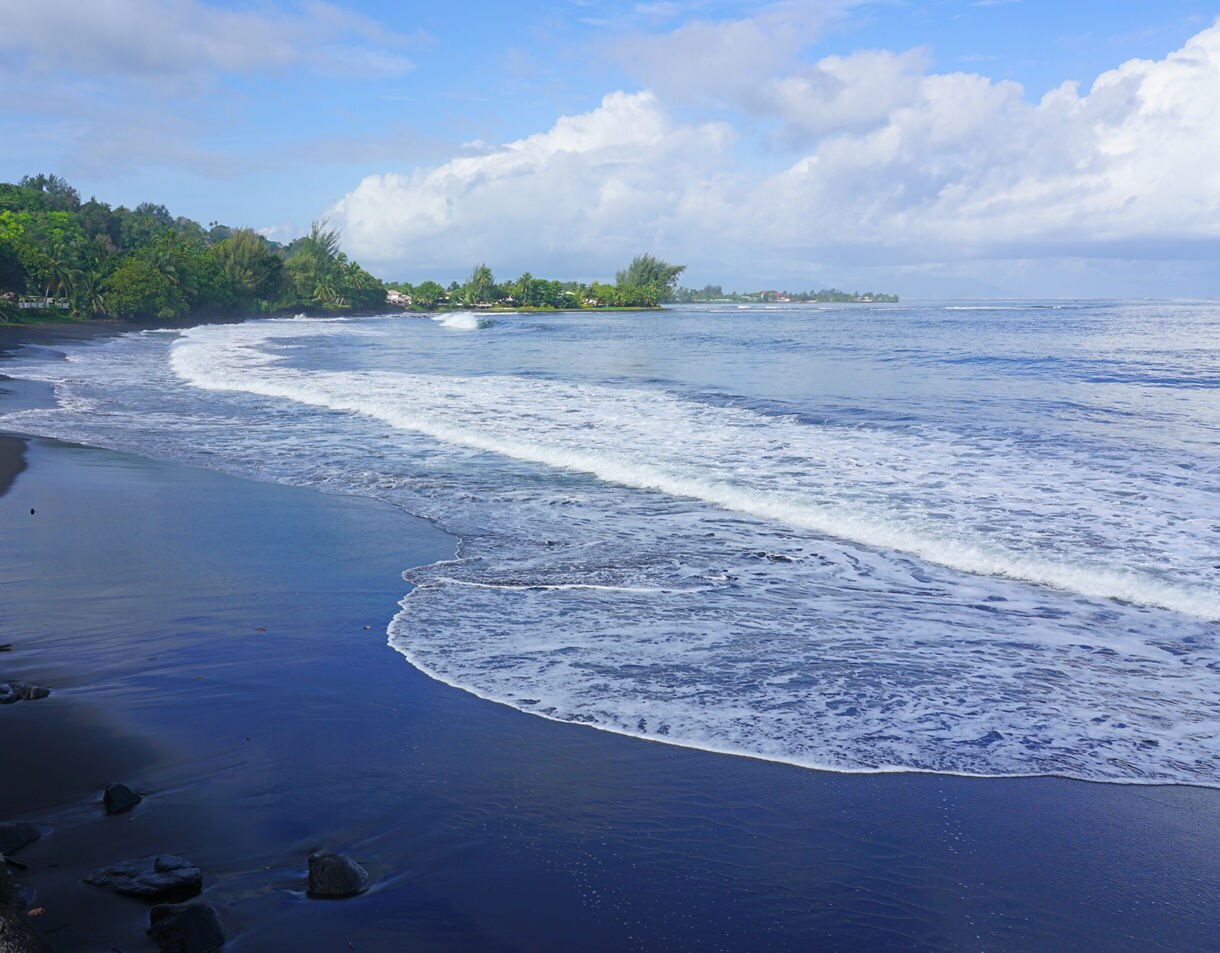 Black sand shoreline at Matavai Bay with gentle waves rolling in, backed by lush green trees and a bright blue sky.