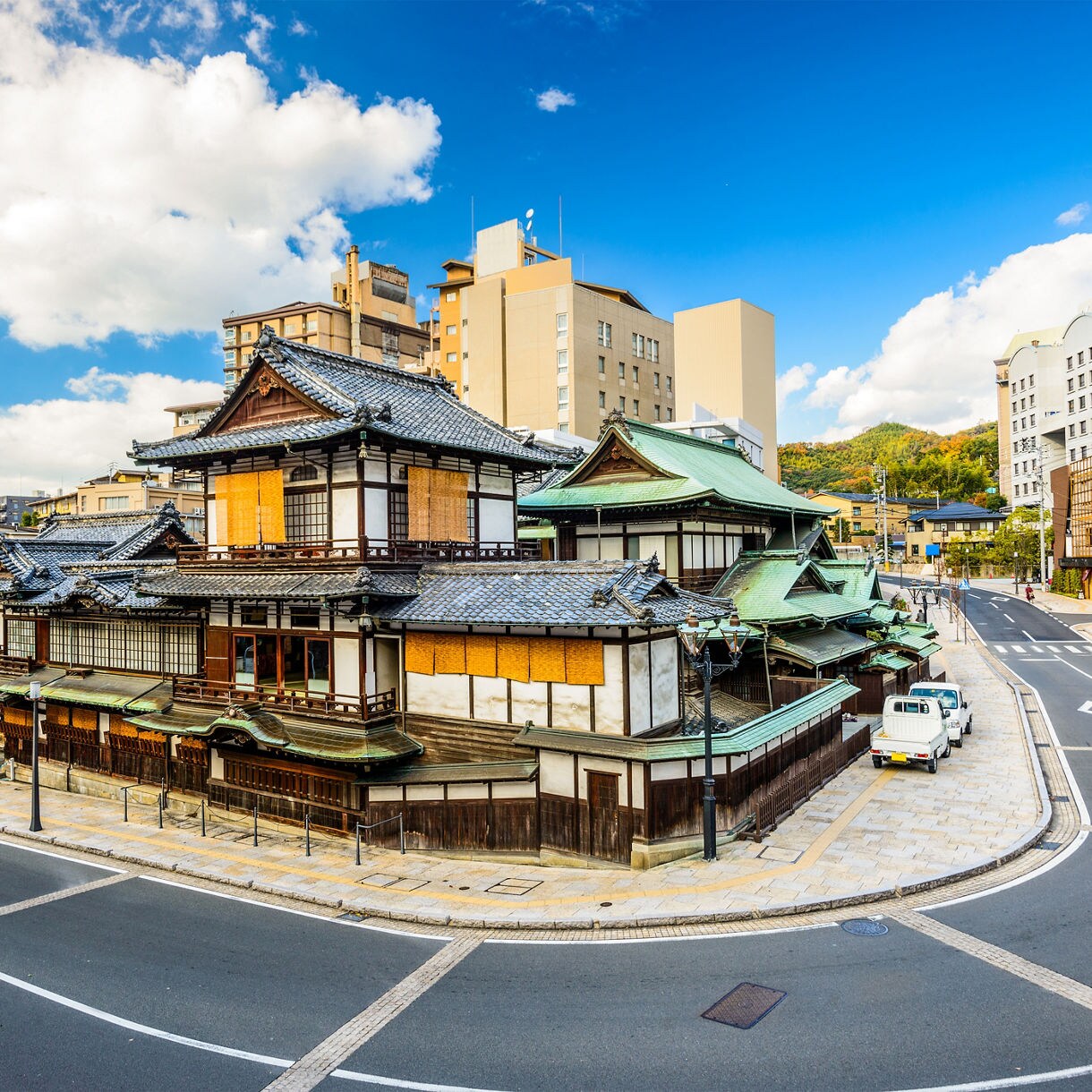 A daytime view of the traditional Dogo Onsen bathhouse with layered tiled roofs and wooden walls, surrounded by modern buildings, wide curving streets and a few parked vehicles under a bright blue sky with scattered clouds.
