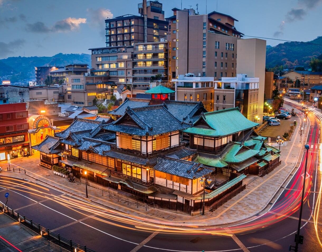 An evening cityscape featuring the traditional Dogo Onsen bathhouse with dark tiled roofs and warm glowing windows, surrounded by modern buildings and streaks of car lights curving through nearby streets under a blue twilight sky.