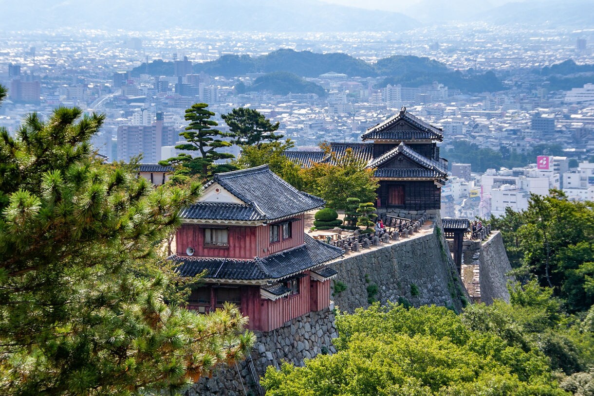 A hilltop section of Matsuyama Castle with traditional wooden buildings and tiled roofs sitting on high stone walls, surrounded by trees, overlooking a dense cityscape of buildings and distant hills.