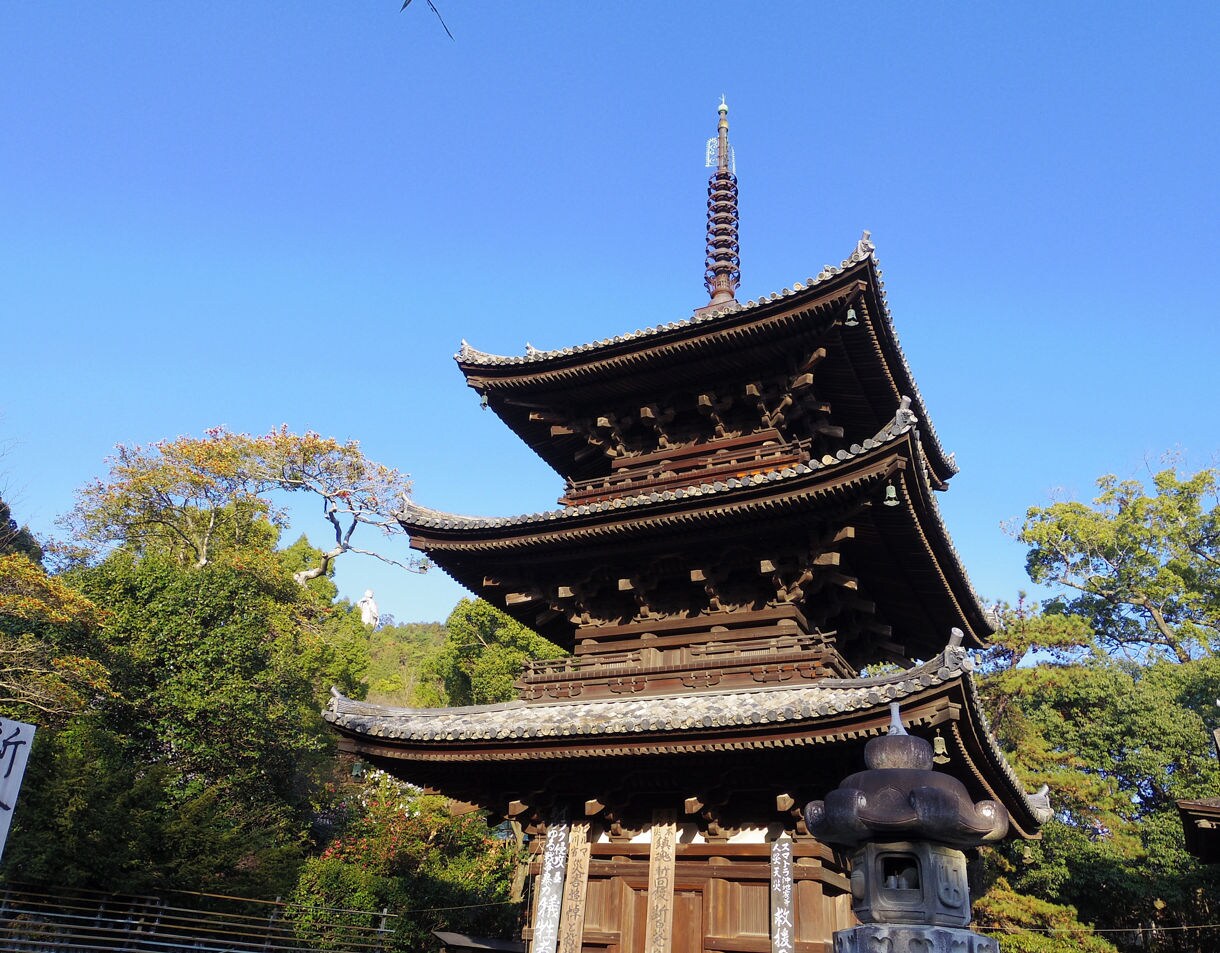 A multi-tiered wooden pagoda with ornate curved eaves and a tall spire, surrounded by green trees and lit by clear afternoon light under a bright blue sky.