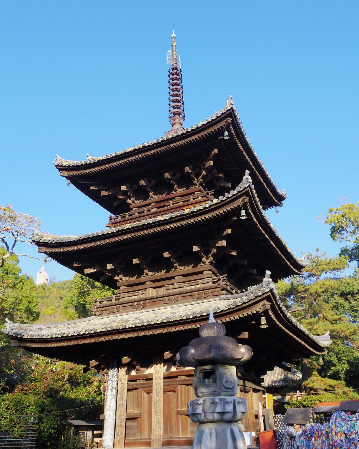 A multi-tiered wooden pagoda at Ishiteji Temple with ornate curved roofs and a tall finial, surrounded by trees and stone lanterns under a clear blue sky.