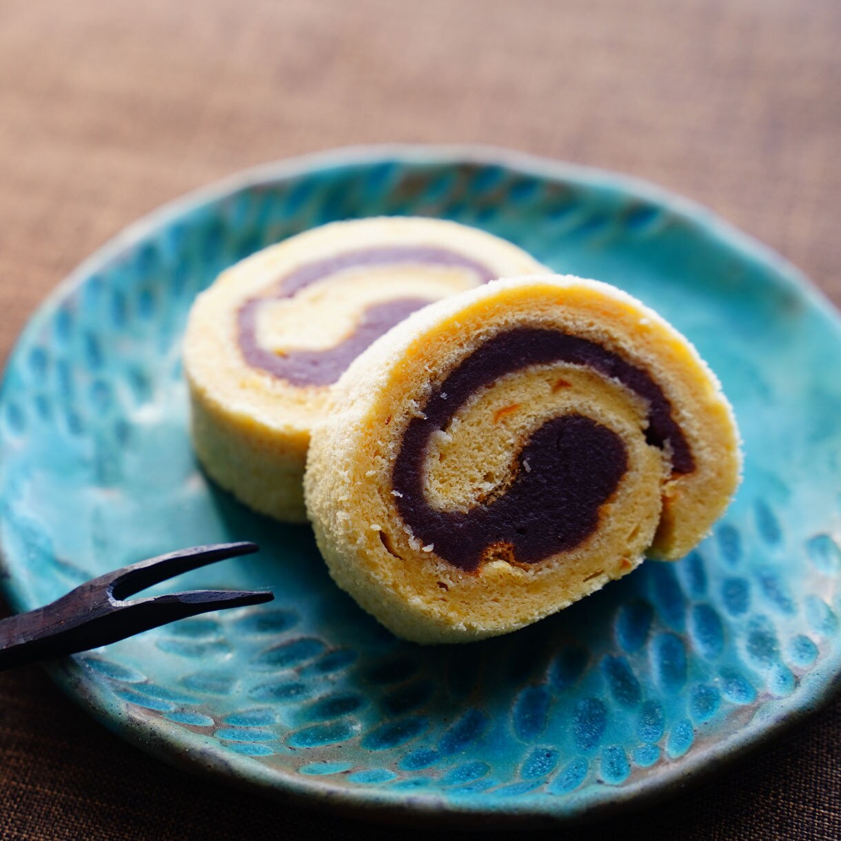 Two slices of Japanese roll cake filled with dark red bean paste sit on a textured turquoise plate beside a small cup of tea and a wooden dessert fork.