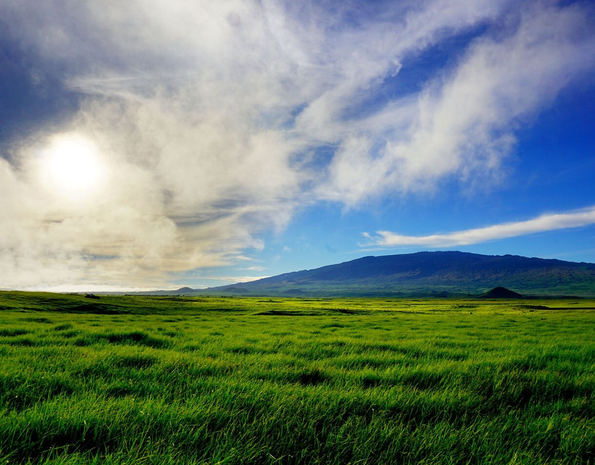 Wide grassy plain leading to the dark outline of Maunakea, with bright sunlight breaking through drifting clouds above.