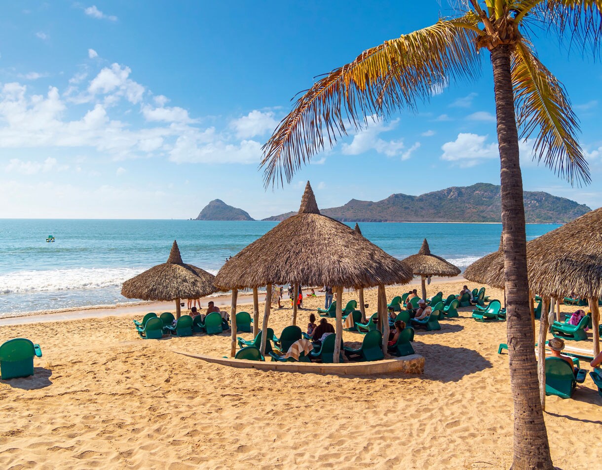 Sandy beach in Mazatlán with palapa huts, lounge chairs, palm trees and distant islands across the calm blue sea.