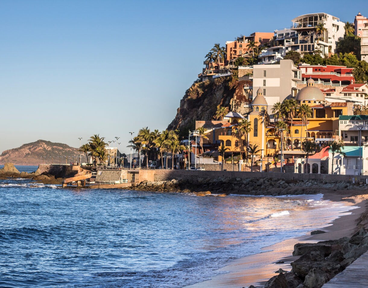 A scenic view of Mazatlán featuring colorful buildings and a prominent yellow church. The setting includes a rocky shoreline and calm ocean waves under a clear blue sky. The image captures the charm of seaside living with a mix of modern and traditional architecture.