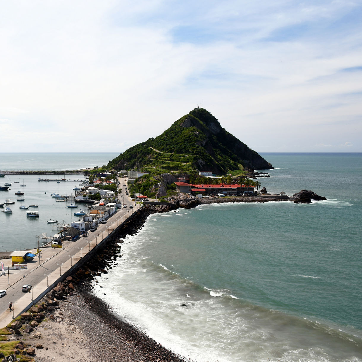 Aerial view of El Crestón Hill in Mazatlán with boats anchored in the harbor, a causeway leading to the hill and waves breaking along the shore.