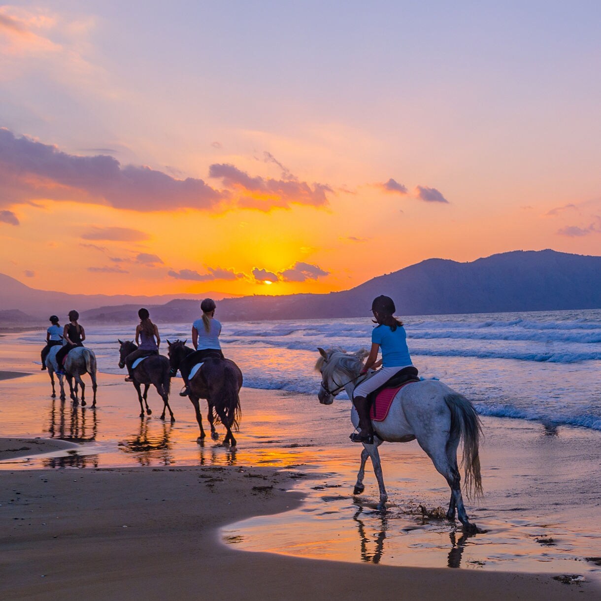 Group of people horseback riding on the beach at sunset with waves rolling in and mountains silhouetted in the background.