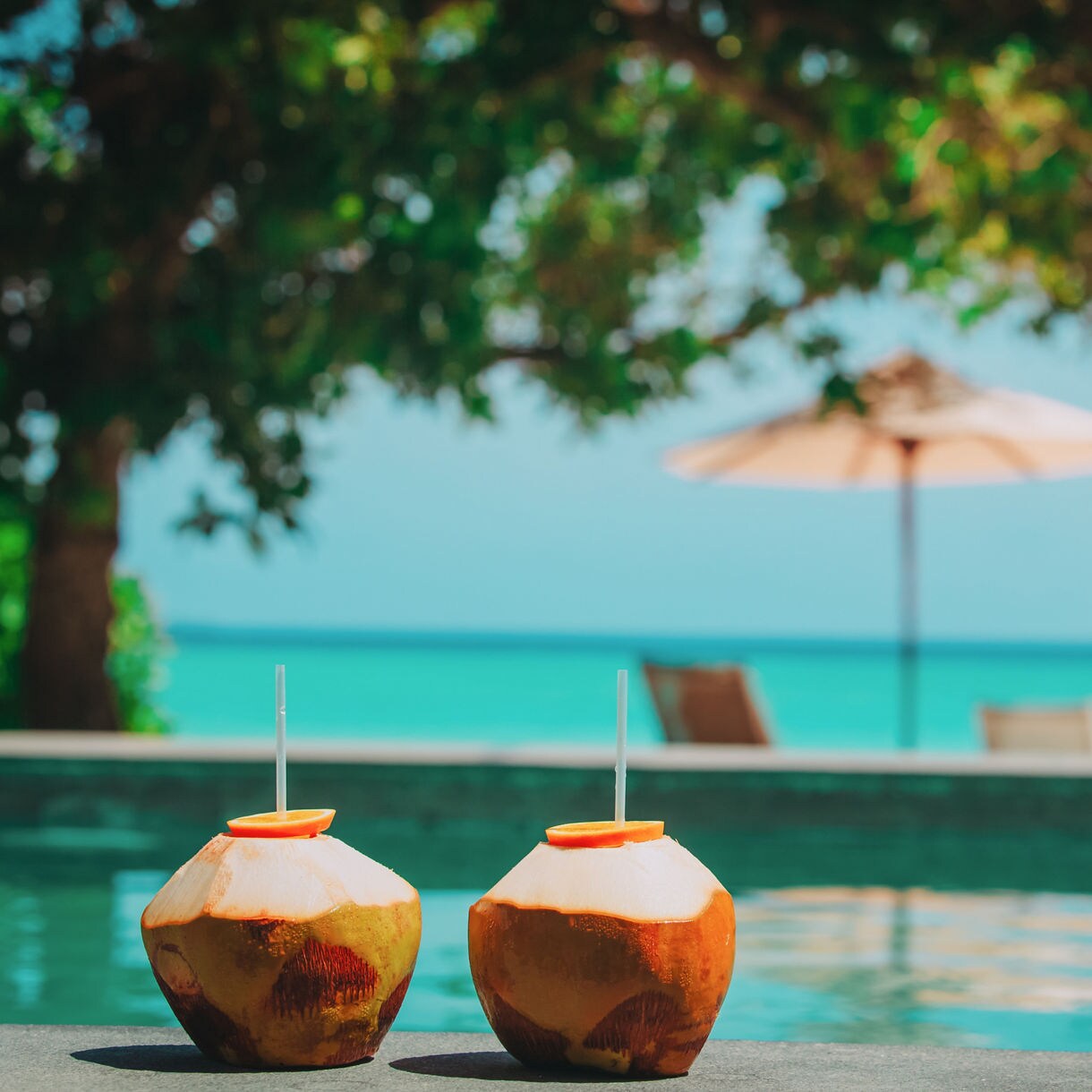 Two coconuts with straws and orange slices set by a pool with lounge chairs, umbrella and ocean view in the background.