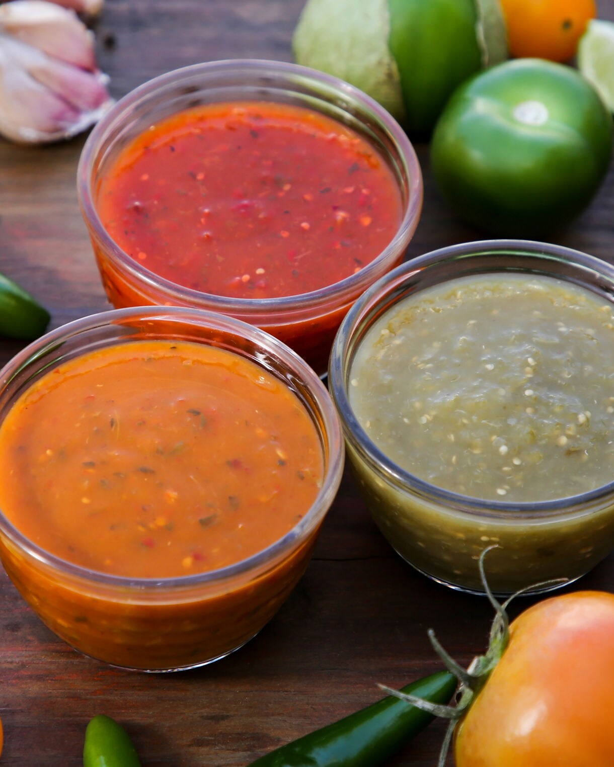 Three bowls of salsa—red, orange and green—surrounded by tomatoes, tomatillos, chili peppers, garlic and lime on a wooden table.