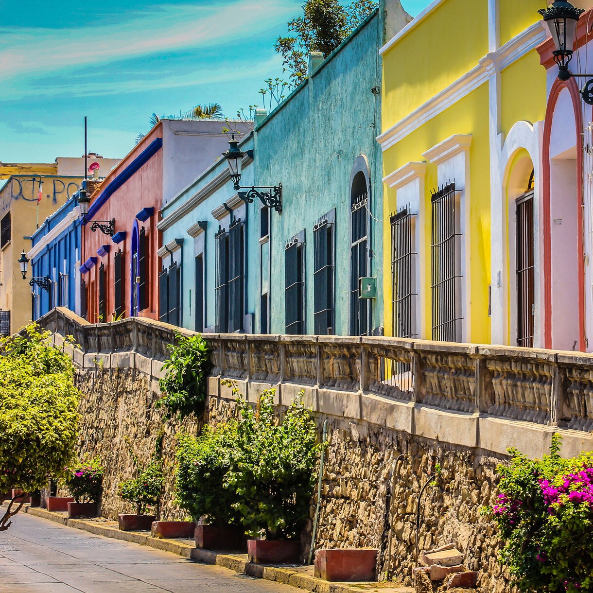 Row of vibrant colonial-style houses in Mazatlán painted in blue, yellow, red and pastel tones, with iron balconies and flowering plants along the sidewalk.