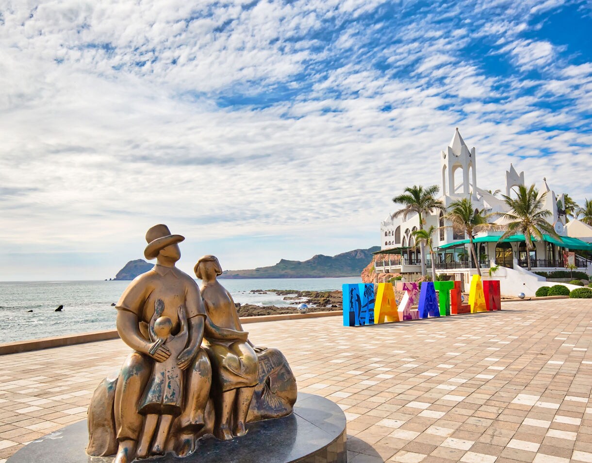 Sculpture of a family on the Mazatlán malecón with the colorful Mazatlán sign, palm trees, and white coastal buildings overlooking the sea.