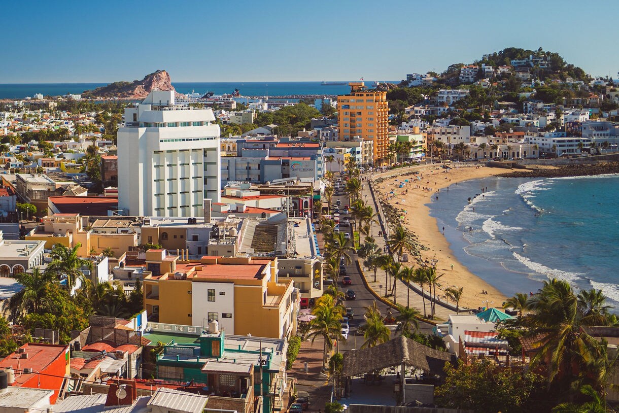 Aerial view of Mazatlán’s beachfront with hotels, palm-lined streets, sandy shores and waves rolling in from the Pacific Ocean.