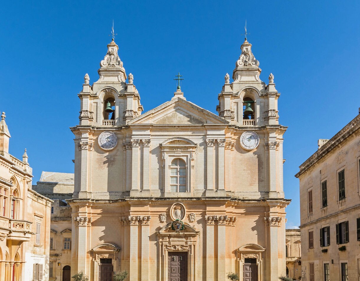Front view of St. Paul’s Cathedral in Mdina, Malta, showcasing twin bell towers, a central entrance and limestone Baroque architecture under a clear blue sky.