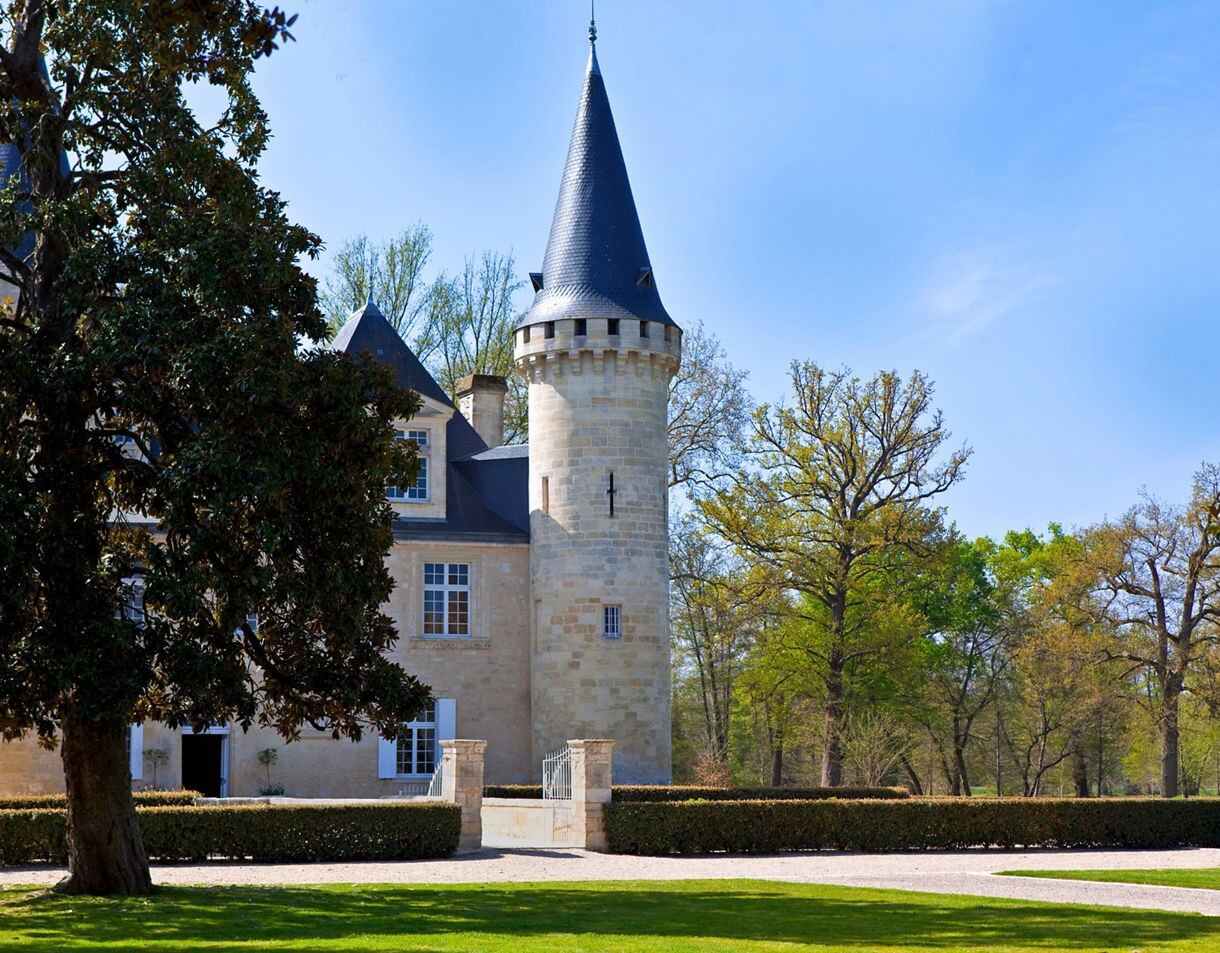  A pale stone château with a pointed turret and slate roof surrounded by manicured hedges and tall trees under a clear blue sky.