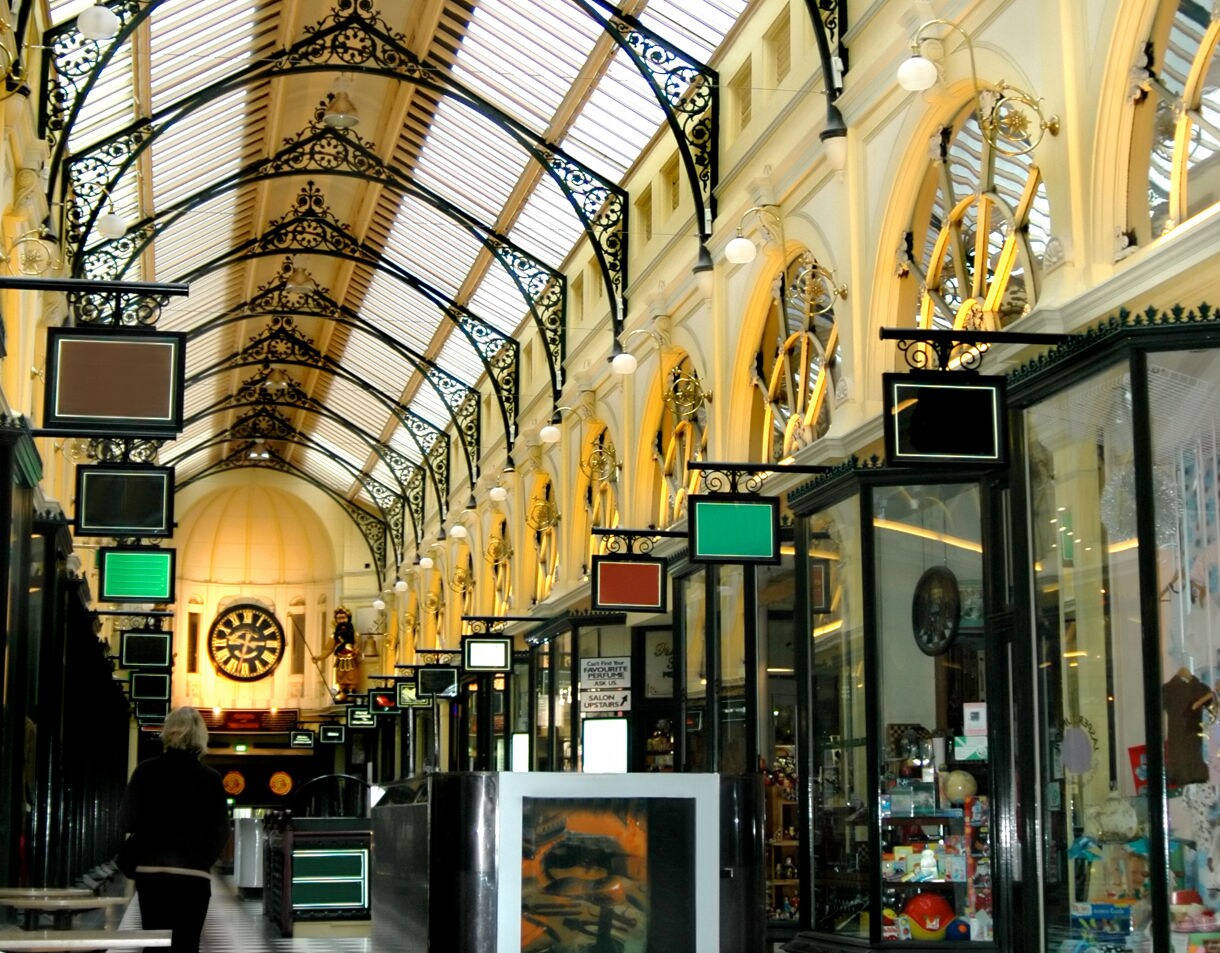 Interior of the Royal Arcade in Melbourne with a black-and-white tiled floor, decorative iron arches, glass storefronts and a large clock at the far end.