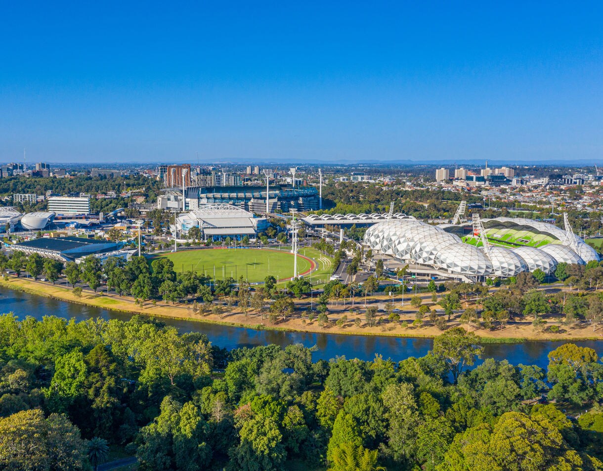 Aerial view of Melbourne’s sports district featuring stadiums, tennis courts and green fields along the Yarra River under a clear blue sky.