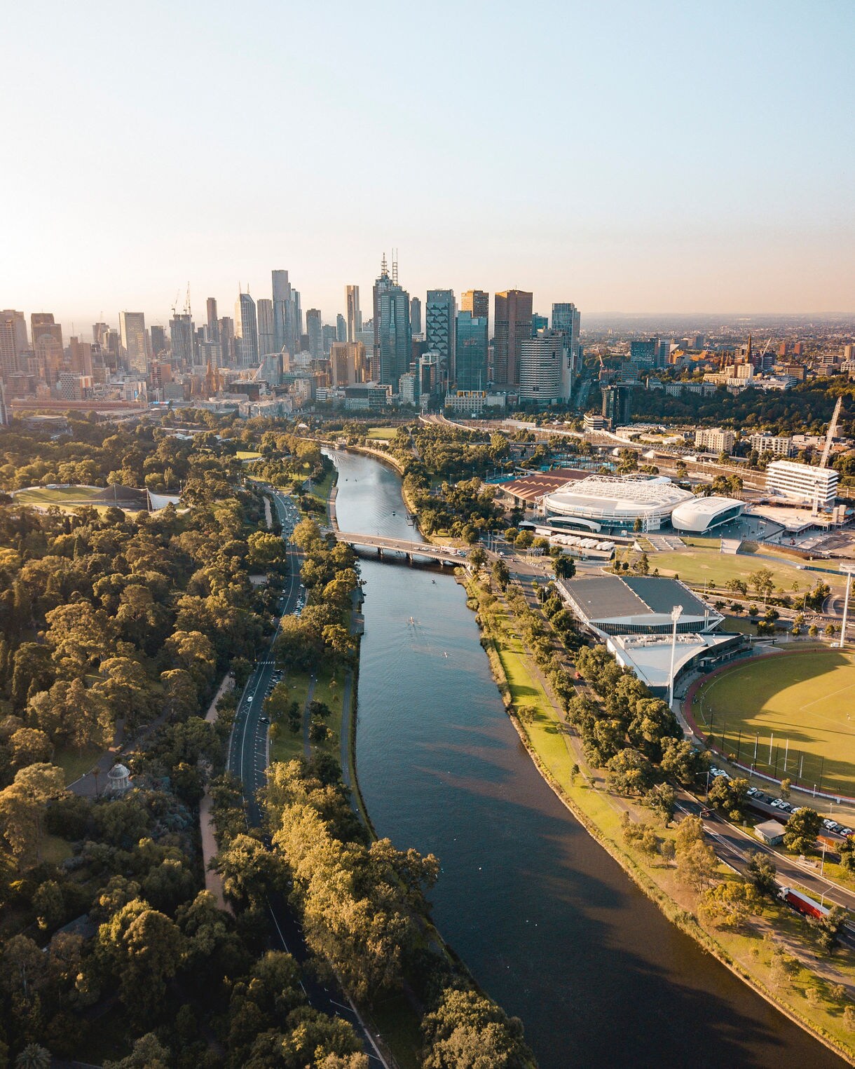 Aerial view of Melbourne with the Yarra River flowing through green parklands, sports stadiums and the central business district under a hazy sky at sunset.