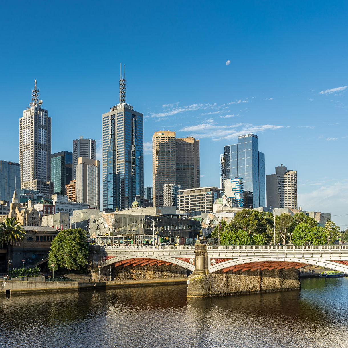 Melbourne skyline seen across the Yarra River with St Paul's Cathedral spires, glass skyscrapers and a stone bridge crossing the water in late afternoon light.