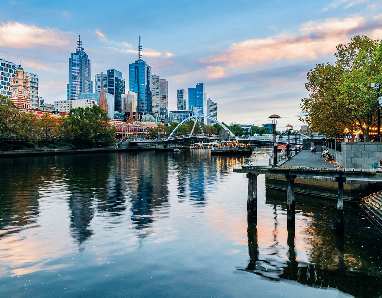Melbourne skyline at dusk with skyscrapers, the Evan Walker footbridge and reflections on the Yarra River, framed by leafy trees and riverside walkways.