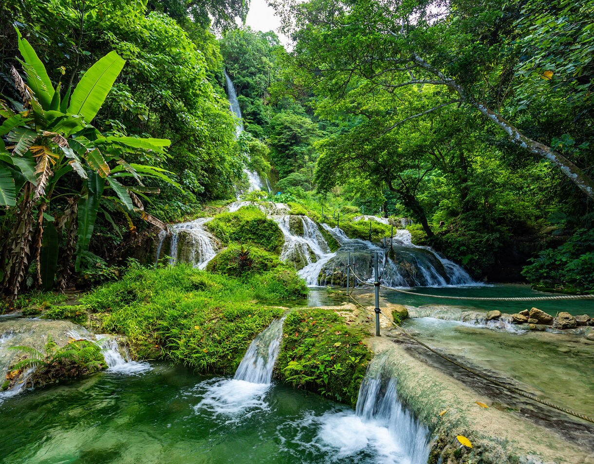 Layered waterfalls spilling over green moss and rocks into clear turquoise pools surrounded by dense tropical foliage.