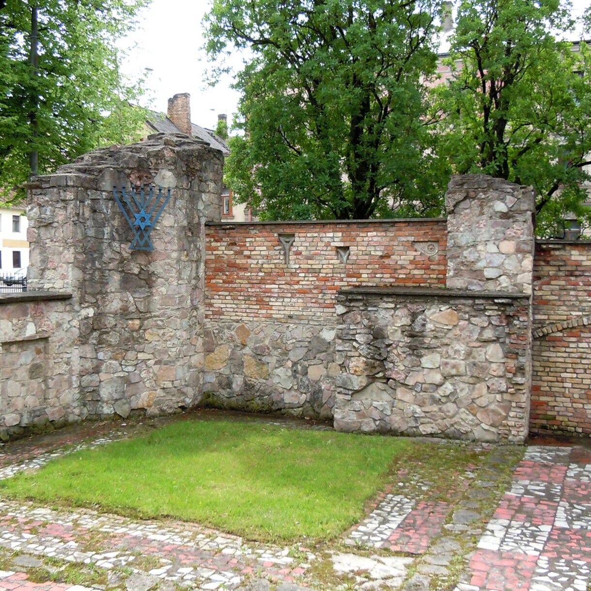 Stone and brick remains of the Great Choral Synagogue in Riga, Latvia, with a memorial symbol on the wall and grass growing within the ruins surrounded by trees.