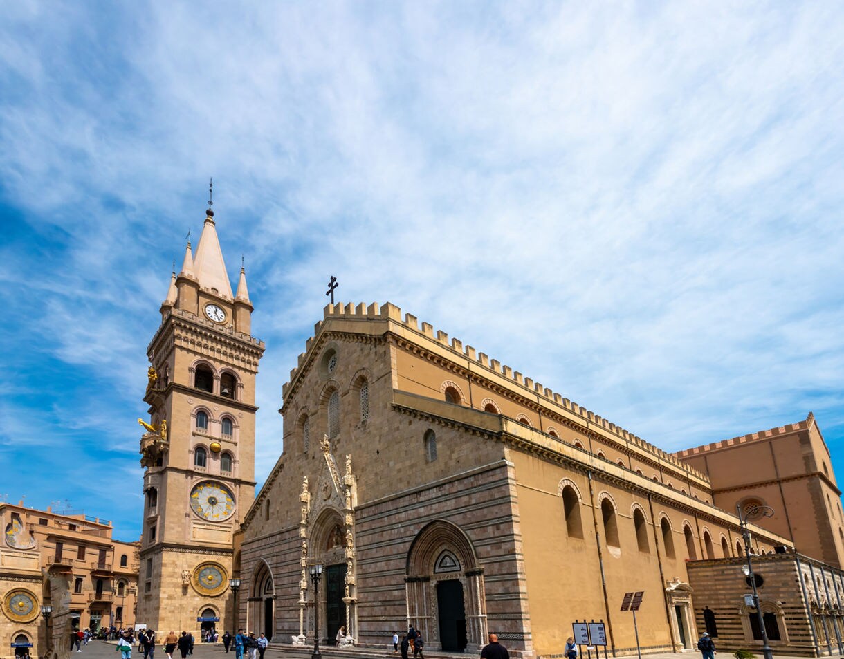 Exterior of Messina Cathedral in Sicily, featuring its tall clock tower with golden figures and ornate astronomical dials, surrounded by visitors under a bright blue sky.
