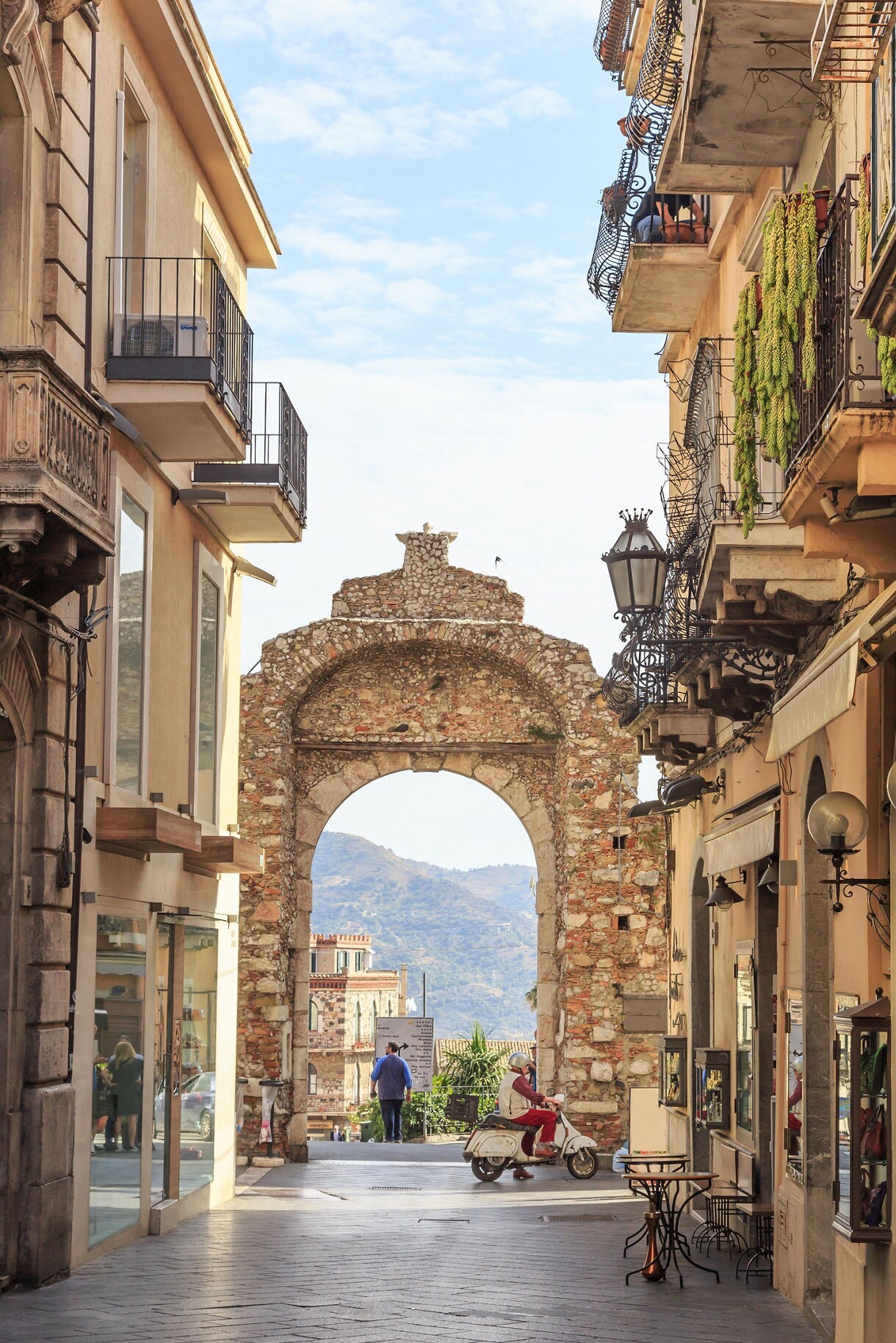 A narrow pedestrian street in Sicily with old balconies and shops leading to a stone archway, beyond which hills and rooftops are visible.