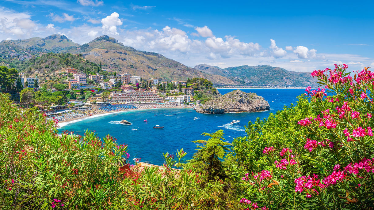 View of Taormina’s coastline with turquoise water, hillside buildings and beaches, framed by pink blooming flowers and green shrubs.