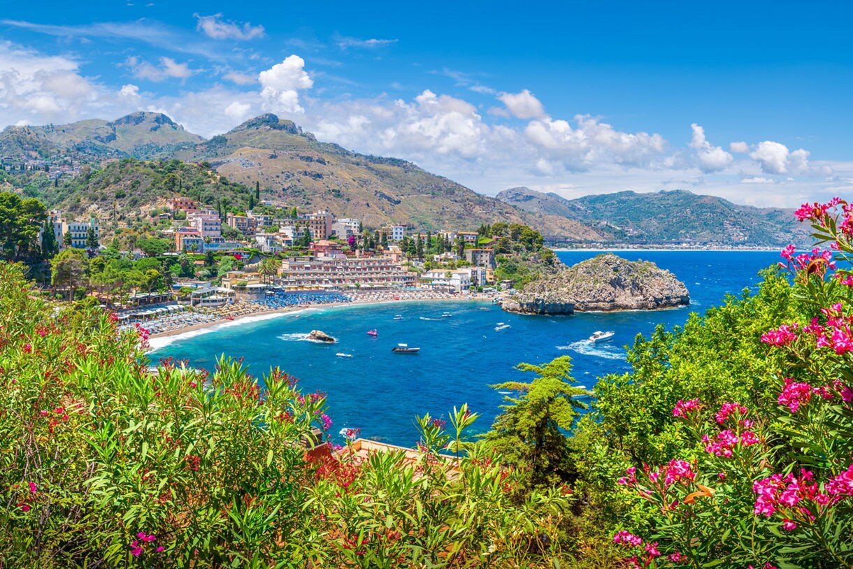 View of Taormina’s coastline with turquoise water, hillside buildings and beaches, framed by pink blooming flowers and green shrubs.
